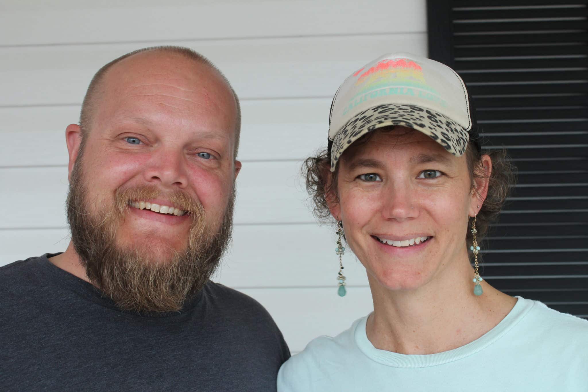 Close up photo of Chris and Julie smiling at the camera at Marriage Getaway. Chris is bald and has a beard while Julie has a baseball cap atop her curly hair and long, dangly turquoise earrings on.