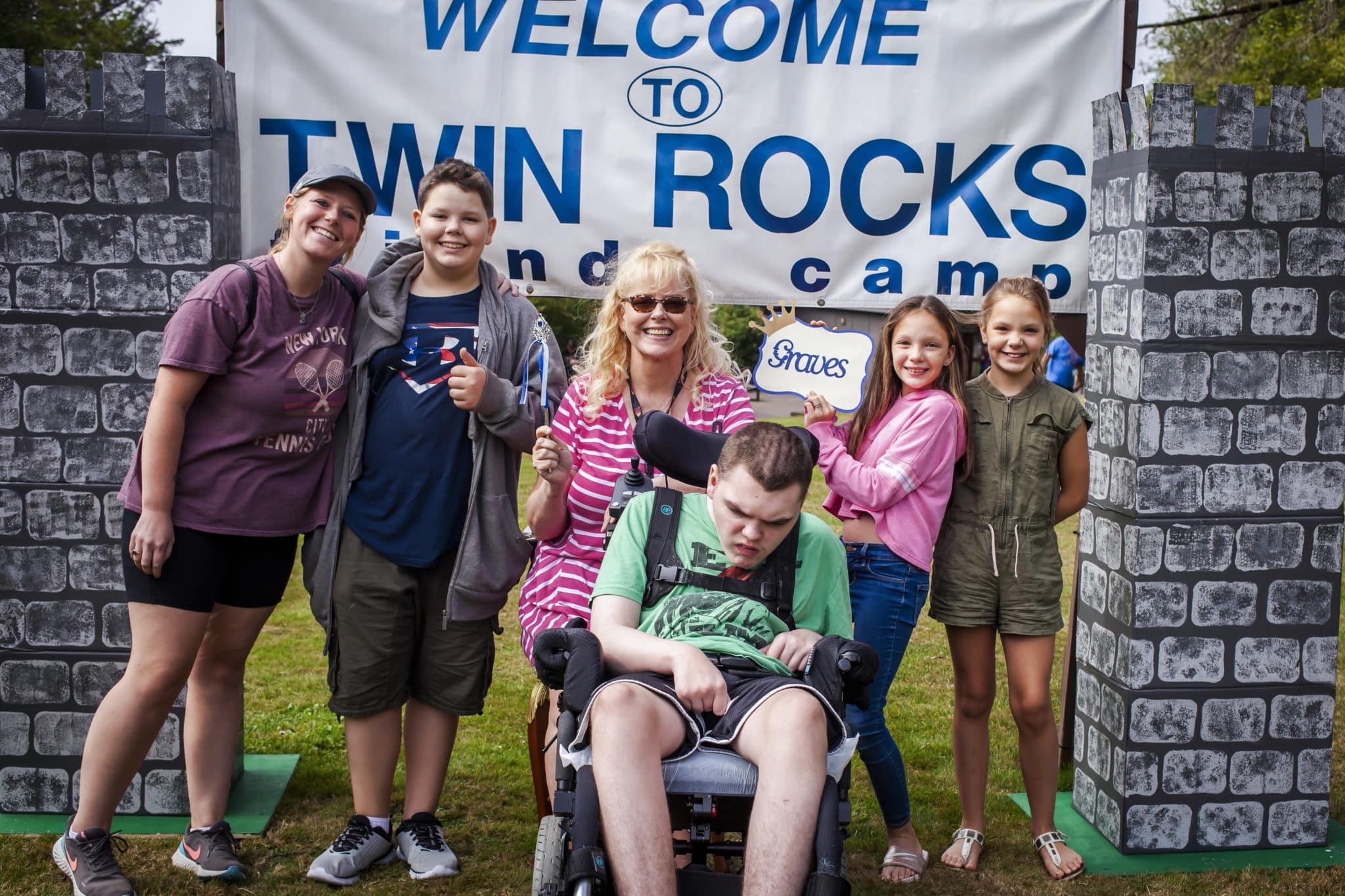 A family portrait of the Shawna, Cody seated in his wheelchair and her four other children smiling at the camera, a sign that says, "Welcome to Twin Rocks Camp" is behind them.
