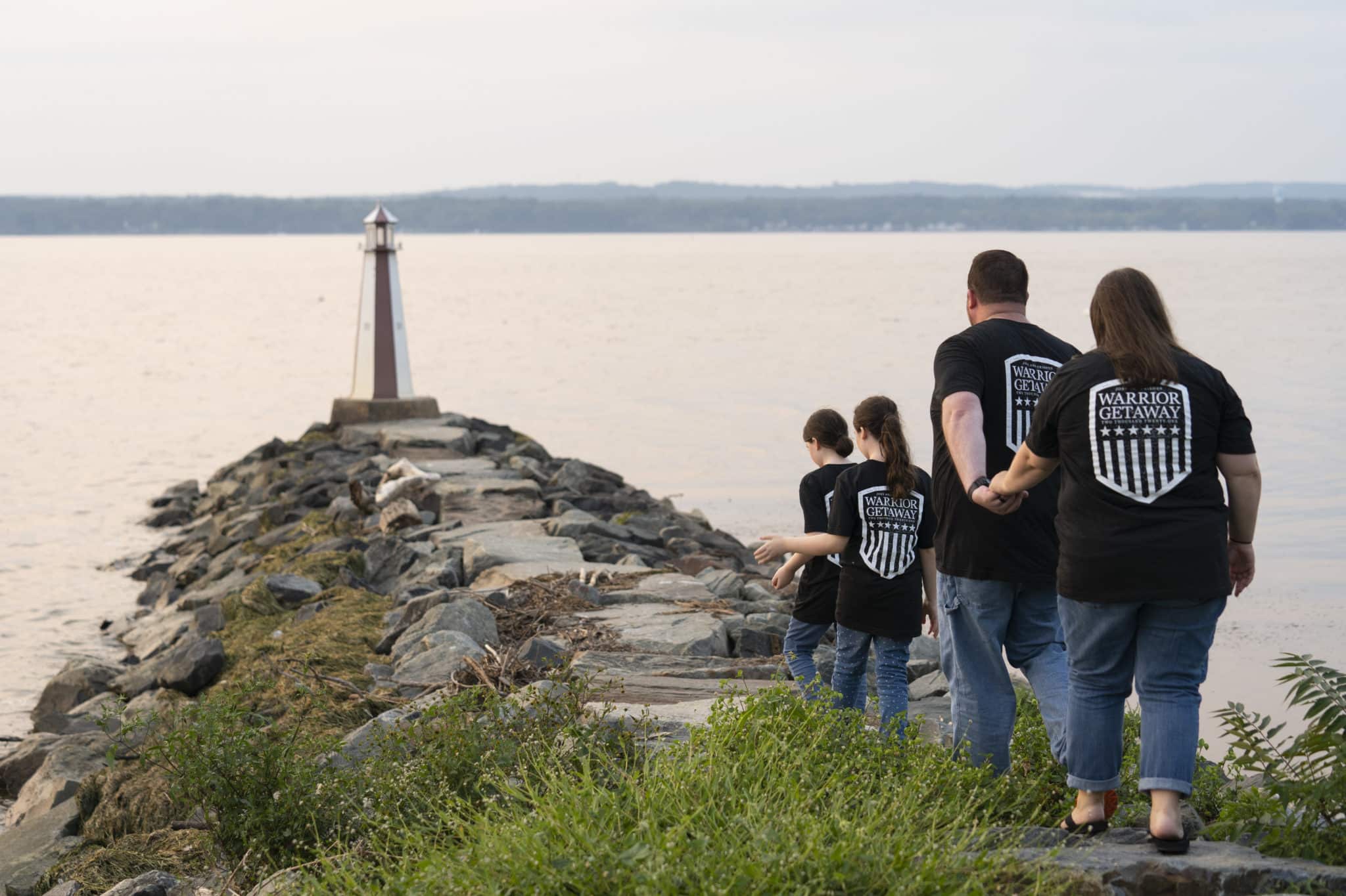 The view from behind a family of four, two children in the front and the parents trailing behind as they walk towards a lighthouse at the end of a stone pathway with a body of water surrounding it.
