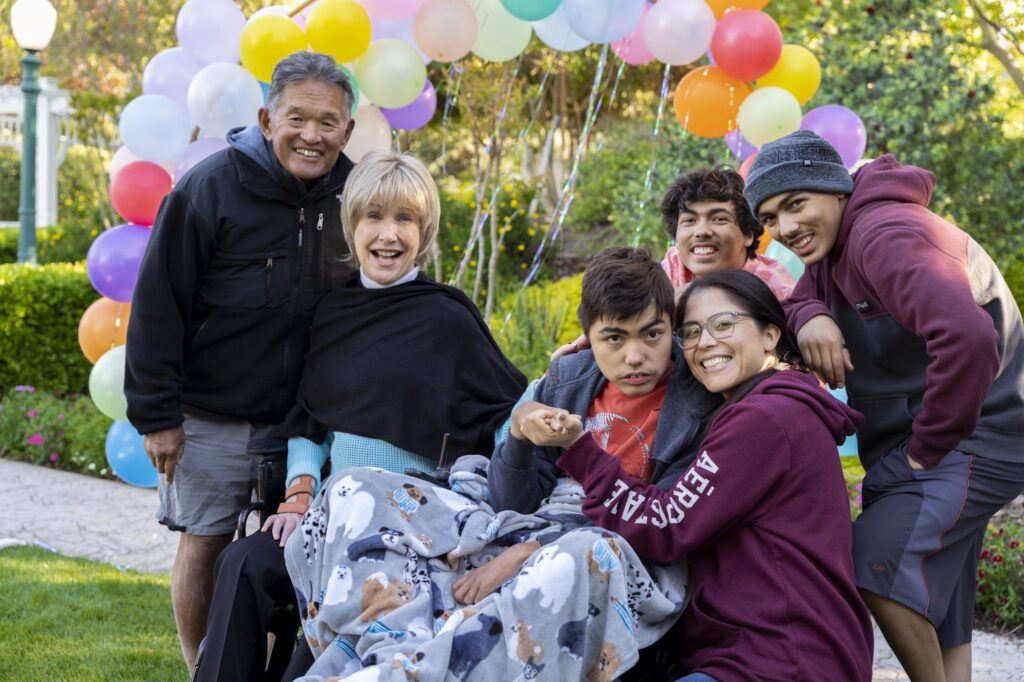 A group photo of Joni and Ken Tada along with Natalia and her four boys, Jeremiah, Izaiah and Nico who is seated in his wheelchair. They're all posing in front of a colorful balloon arch with gardens behind it.