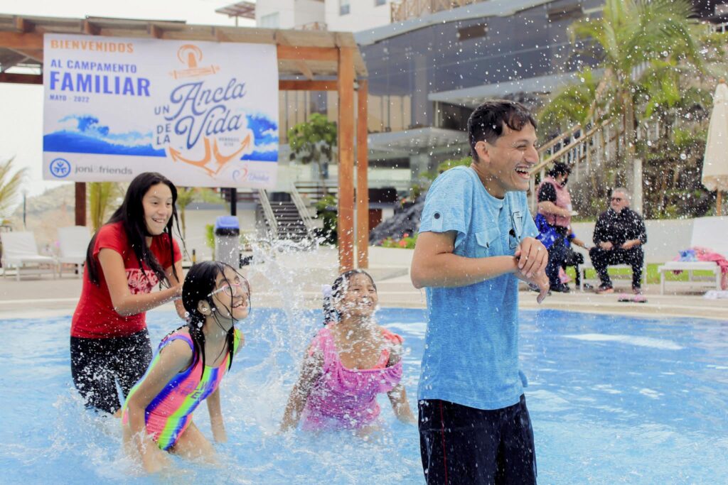 A scene of an older boy in a pool smiling and laughing as three younger girls splash him from behind, a Joni and Friends Family Retreat sign in the Spanish hung in the background.