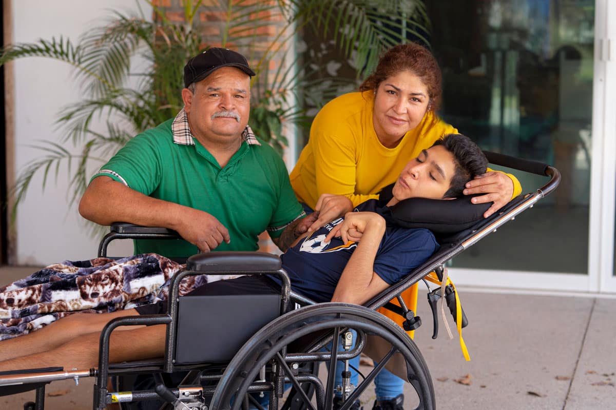 David's parents Leandro and Josefina standing next to him as he's in his wheelchair, looking at the camera.