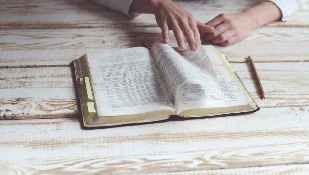 An open Bible on a wooden table, someone flipping through the highlighted pages, a pencil sitting next to it.