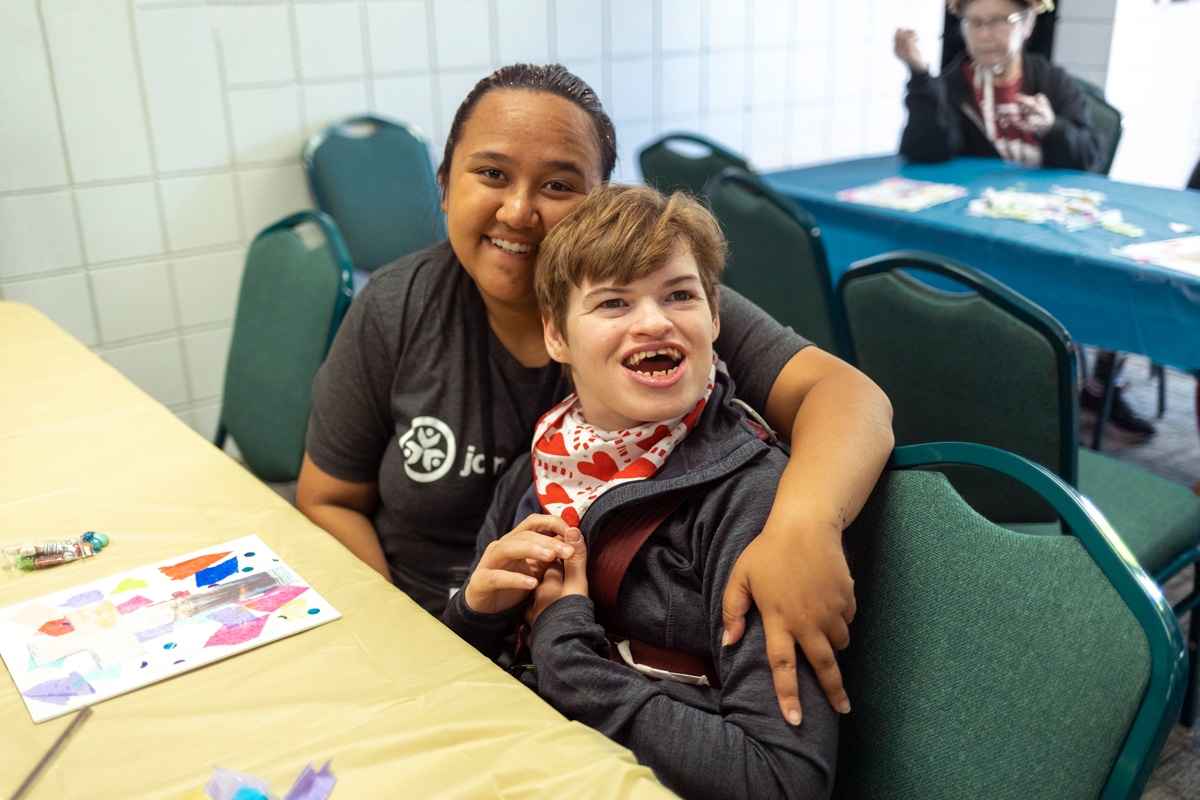 A Joni and Friends volunteer smiling at the camera as she puts her arm around her buddy, a young woman who appears to have a disability, who is also smiling at the camera.