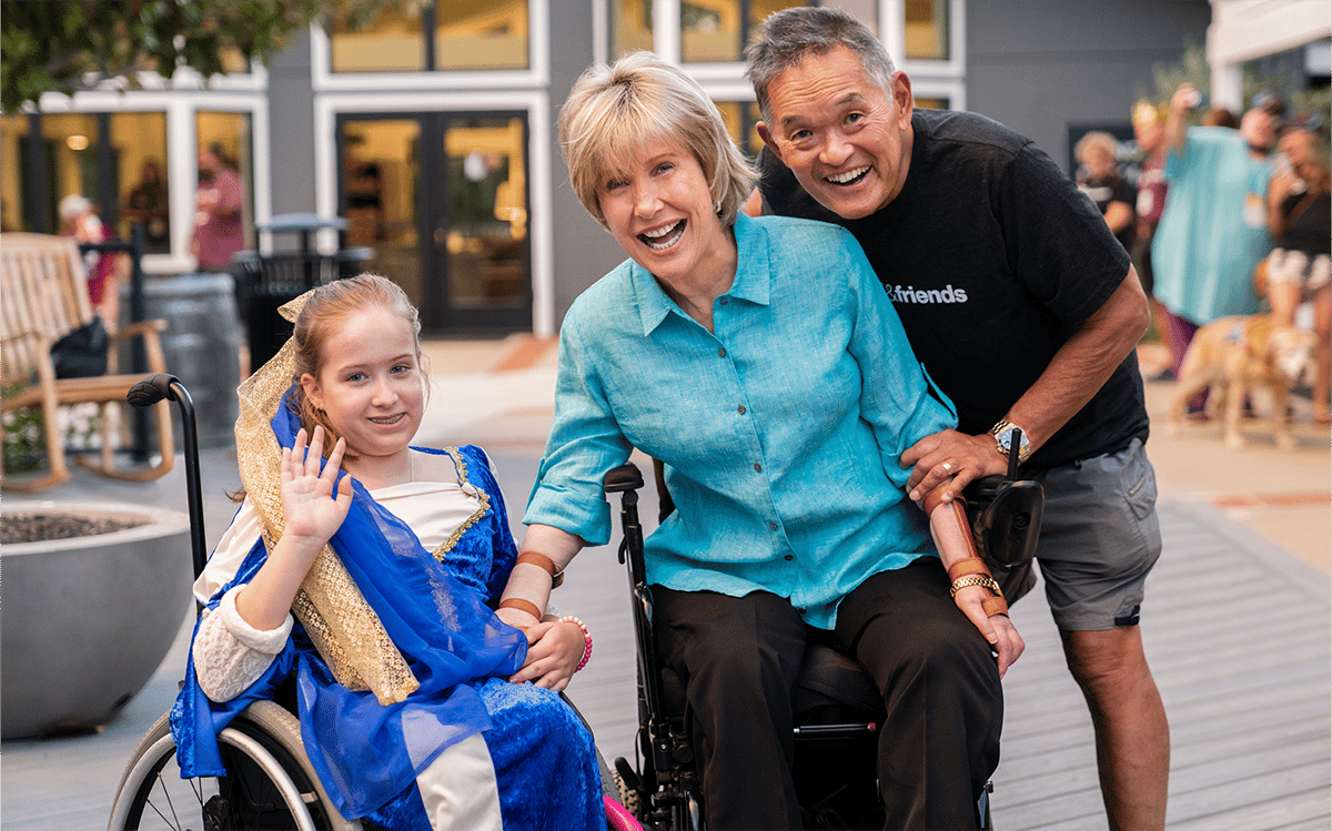 A picture of Joni and Ken smiling at the camera next to a young girl in a princess costume seated in a wheelchair.