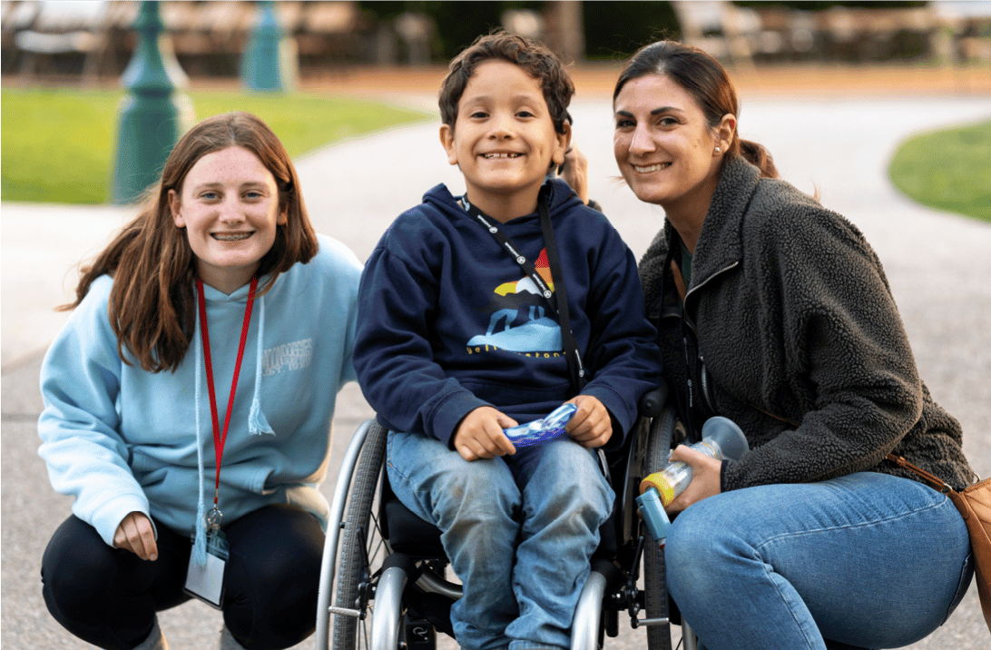 A Joni and Friends volunteer leaning down next to a young boy in a wheelchair, his mother on the other side of him, all smiling at the camera.
