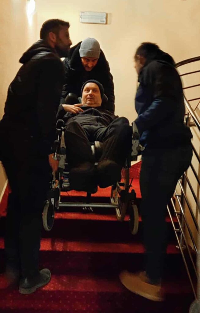 Three people carrying a man in his wheelchair down the stairs.
