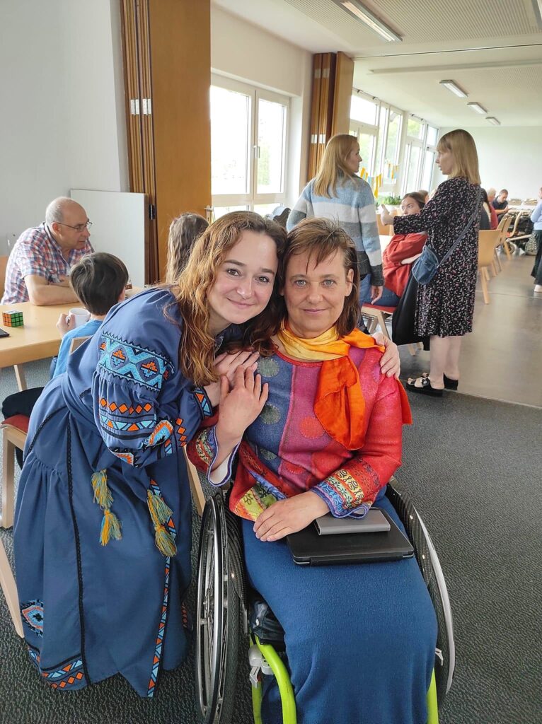 Galyna hugging Martina from the side as she's seated in her wheelchair. Both are smiling at the camera.
