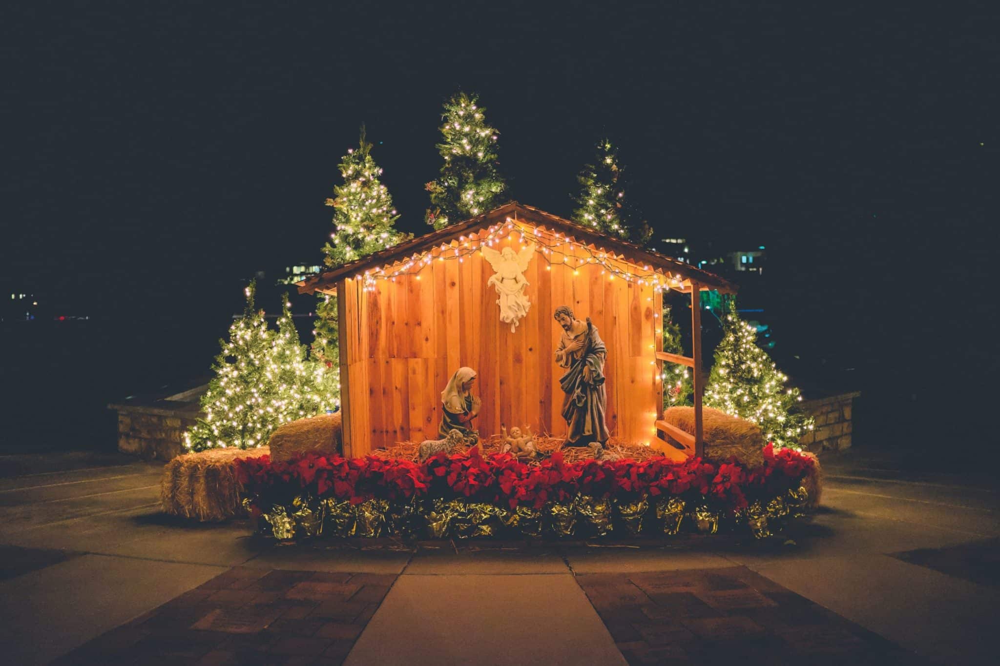 Close up of a wooden nativity scene with Christmas trees in the background behind it and lights all around.