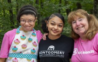A picture of three women with their arms around each other and smiling at the camera. The one on the far left is a young girl wearing a cupcake apron, the girl in the middle is wearing a black college t-shirt and the woman on the right it wearing a pink Joni and Friends shirt.