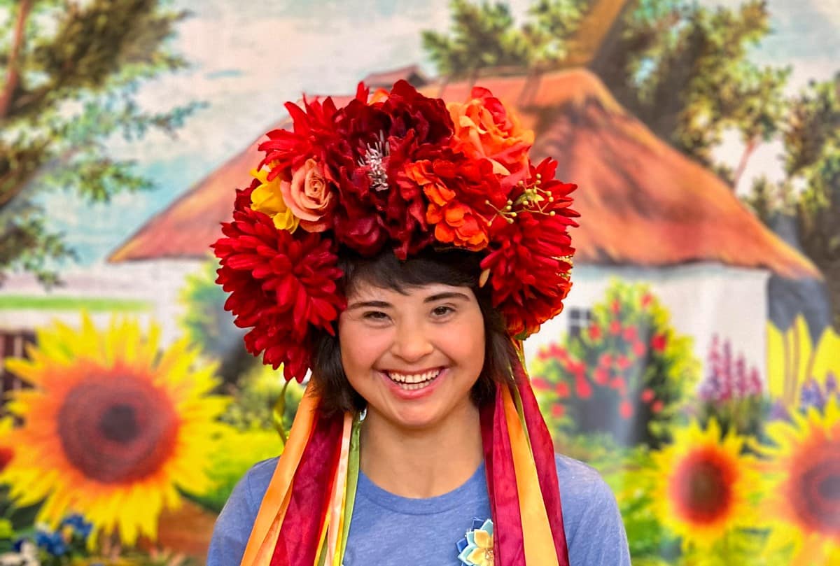Very happy smiling young lady wearing a large flower crown and a Joni friends tee, sunflowers in background.