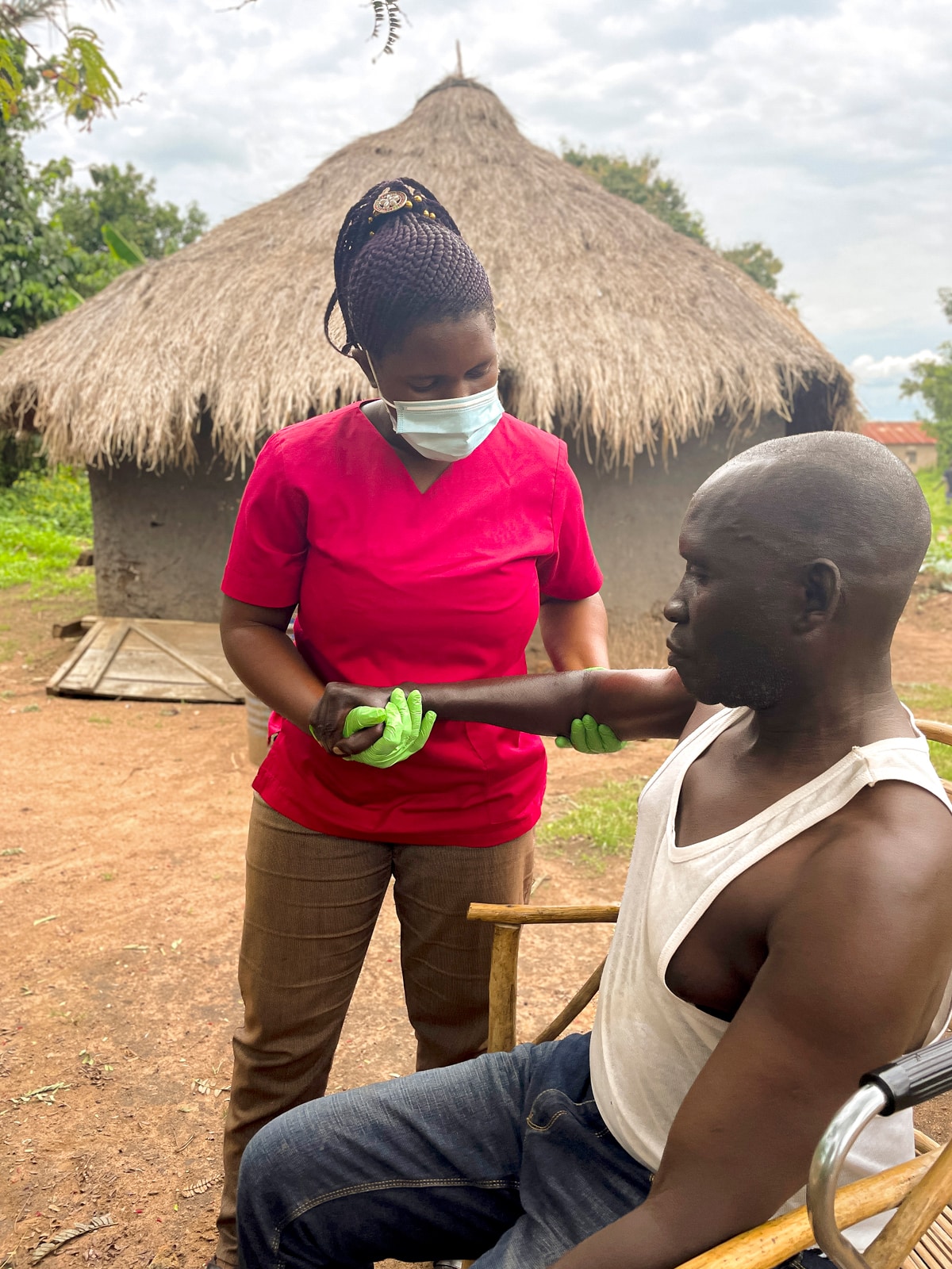 Daisy working on Ashraf's arm as he's sitting in a chair, a small hey-covered hut in the background.