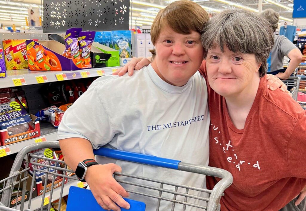 A picture of two women, one younger and one older who appear to have down-syndrome, smiling at the camera and shopping at the grocery store.