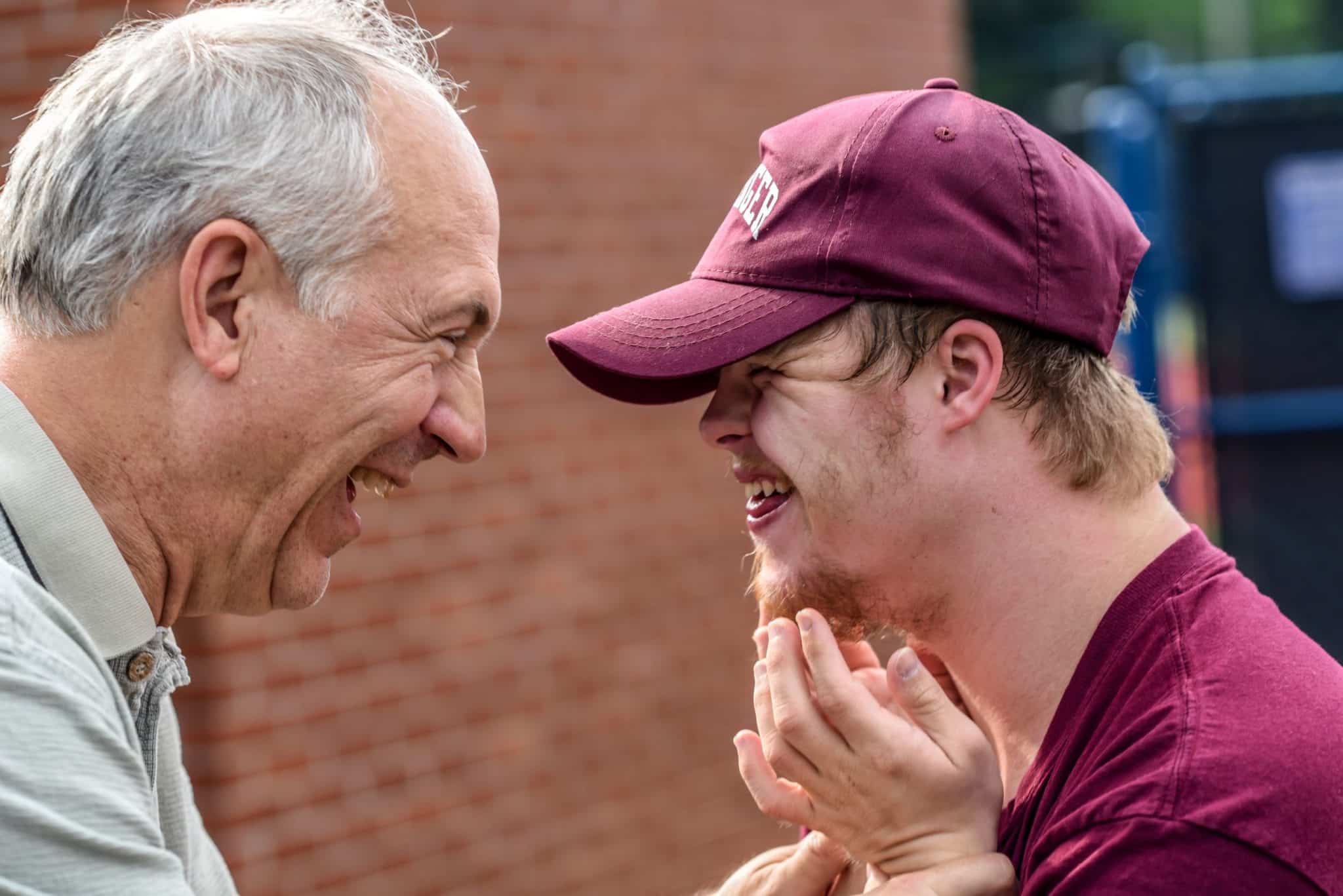 An elderly man smiling and laughing with a young man who appears to have down-syndrome.