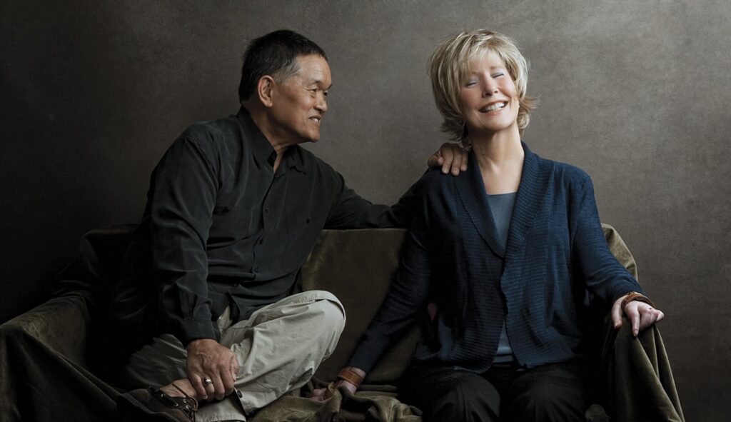 Ken smiling at Joni with his hand on her shoulder as she smiles with her eyes closed. A gray backdrop behind them.