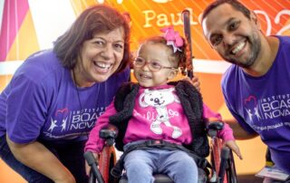 A little girl seated in a wheelchair smiling wide as two Joni and Friends volunteers crouch on either side of her, smiling at the camera.