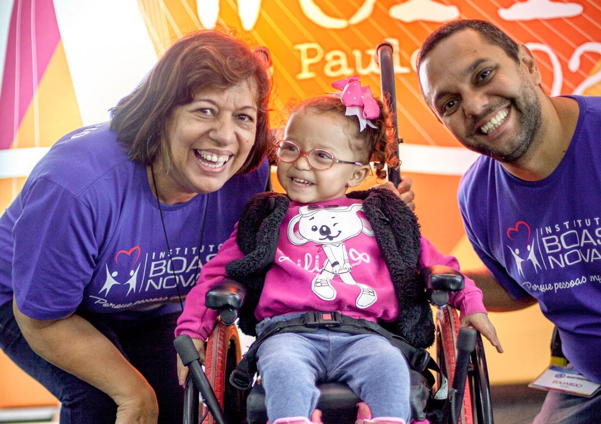 A little girl seated in a wheelchair smiling wide as two Joni and Friends volunteers crouch on either side of her, smiling at the camera.