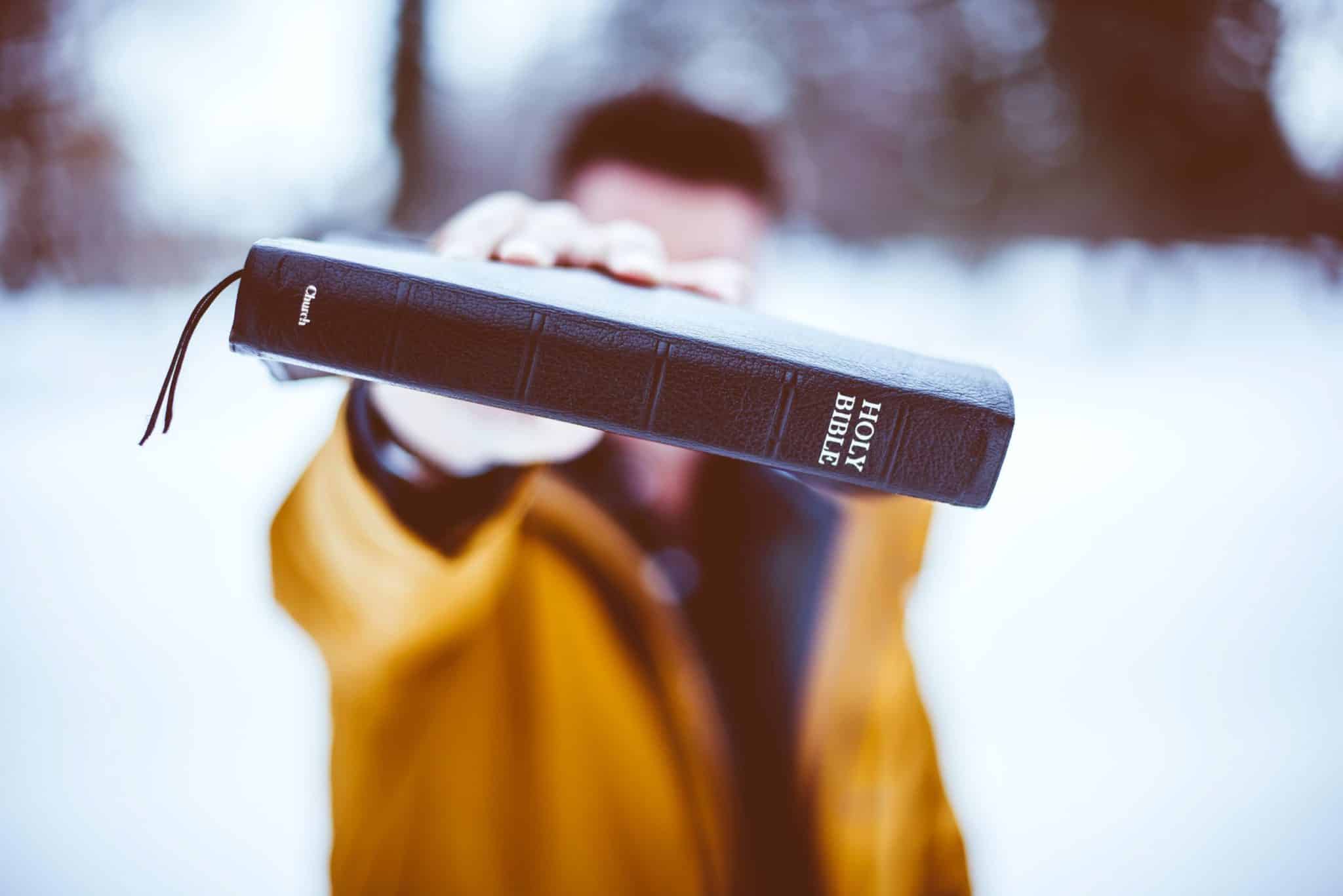 A man in a snowy environment holding out a bible sideways so the words "Holy Bible" are clearly seen.