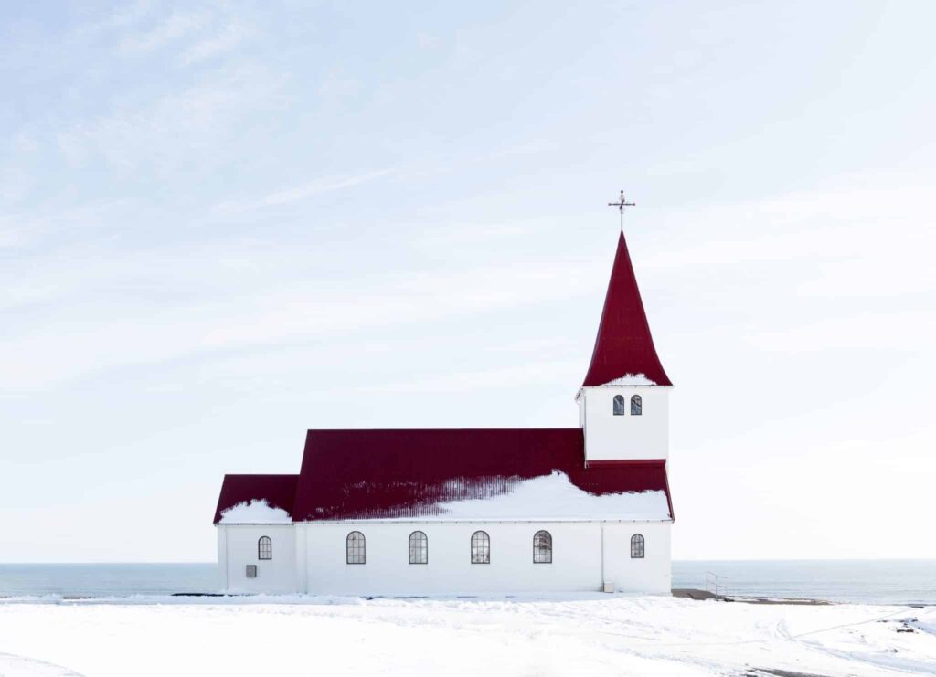 A church with a red roof, snow surrounding it and a large body of water beside it.