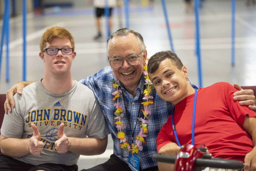 An elderly man with a Hawaiian lei on with his arm around a young man with Down-Syndrome on his left a young man using a mobility device on his right, all smiling at the camera.