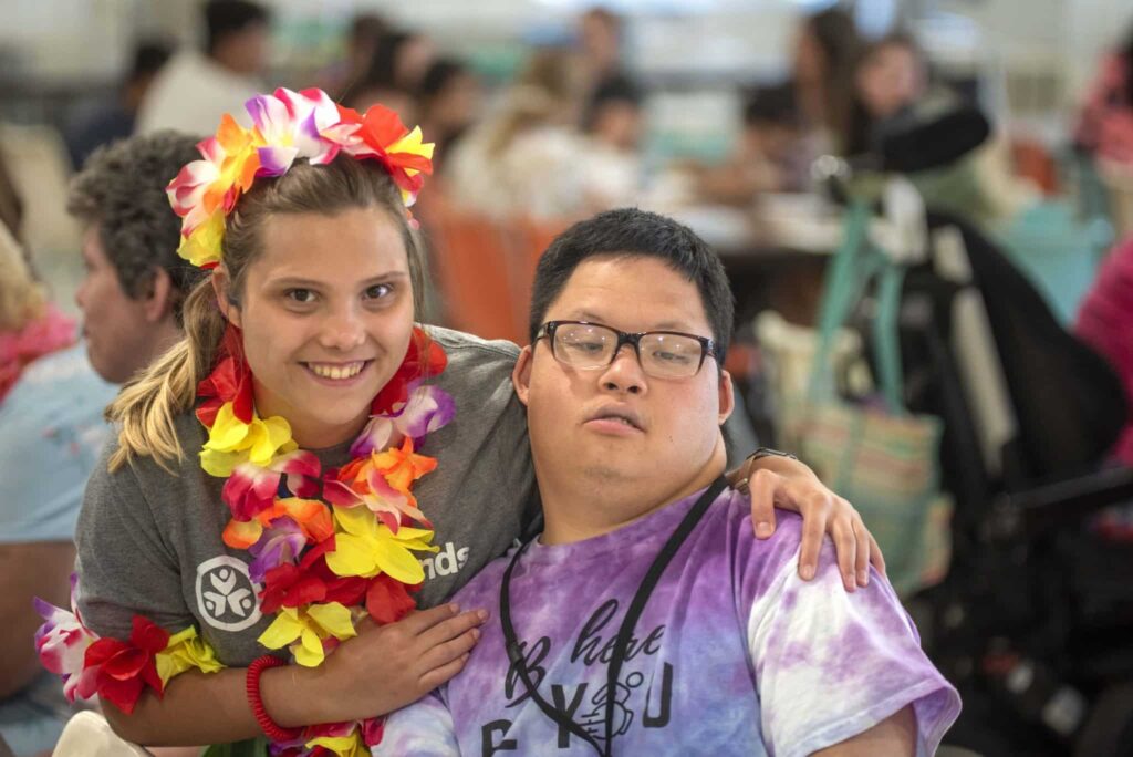 A young woman with a brightly-colored Hawaiian lei on side-hugging a young man with Down Syndrome.