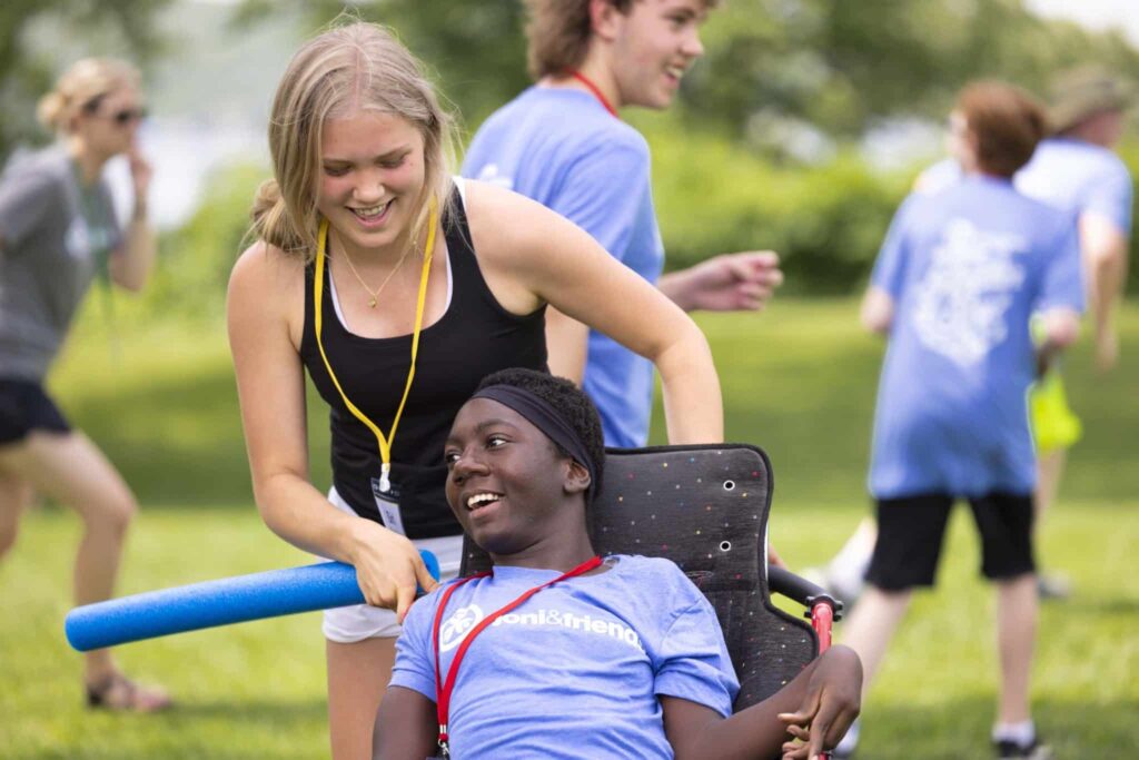 A young blonde girl pushing a young African-American girl in a wheelchair around a grass field as they play a game.