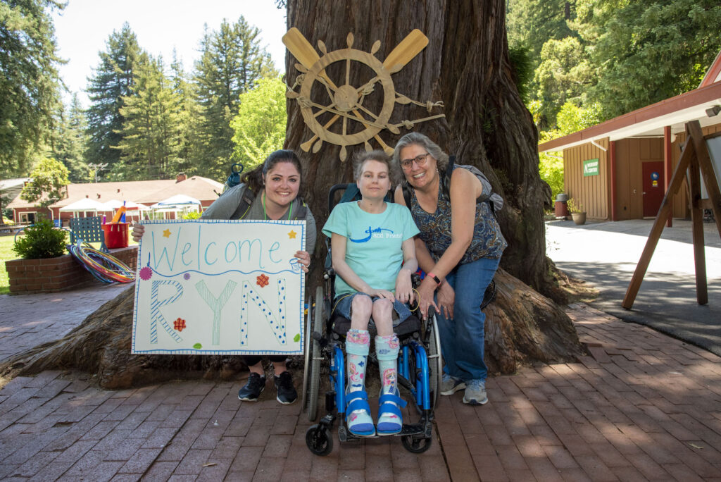 Deanna leaning down next to Cathryn who is seated in her wheelchair, a Joni and Friends volunteer next to them with a sign that says "Welcome Ryn." All are posing in front of the trunk of a large tree and smiling at the camera.