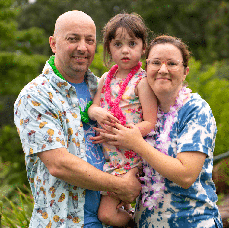 A group photo of a mother and father as they hold their small daughter who has autism in the center of them. They're all wearing Hawaiian leis and smiling at the camera.