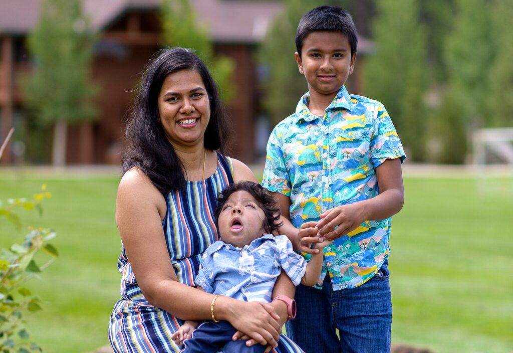 A mother carrying her son, who has a disability, while her other son stands beside them.