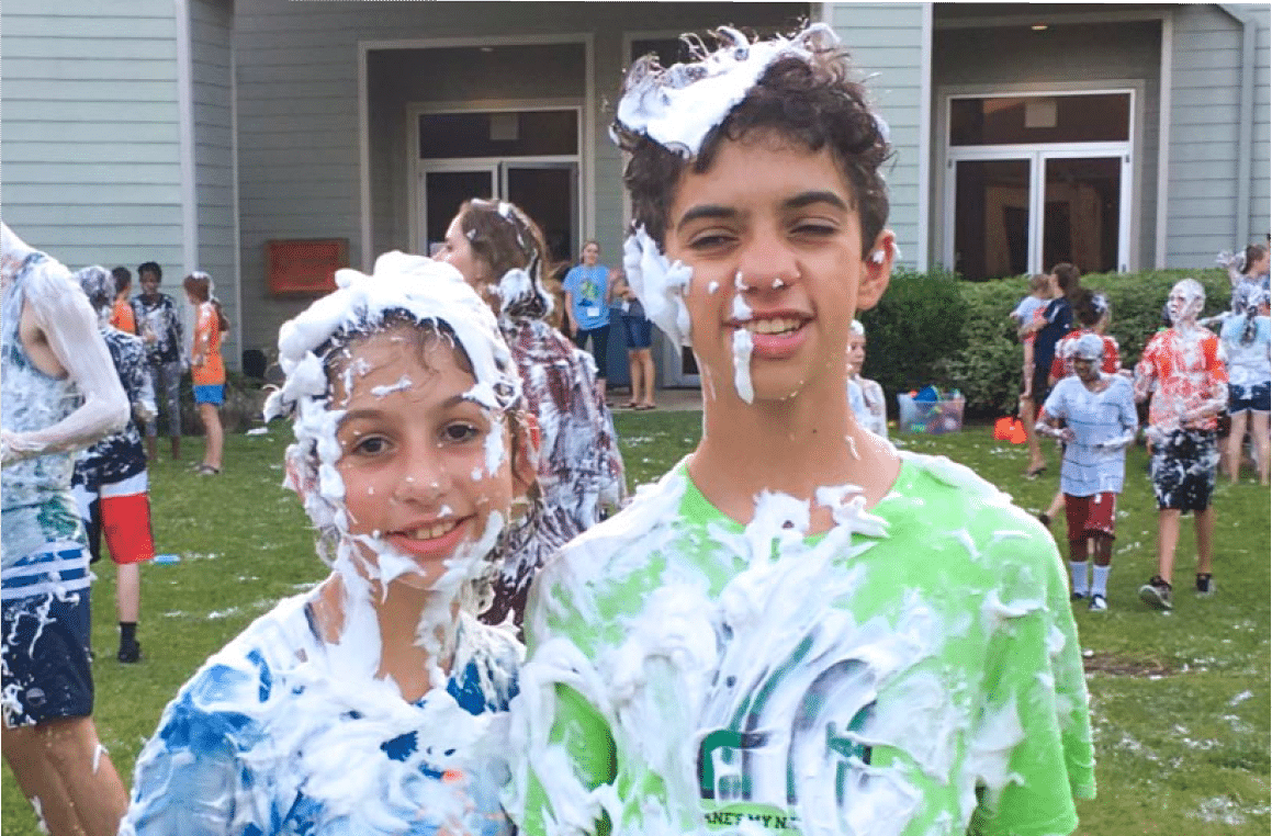 A little girl and little boy with shaving cream all over them and smiling at the camera.