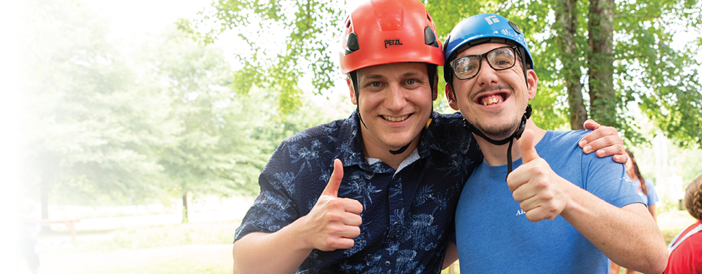 Two men wearing protective helmets, smiling and giving a thumbs up, with lush greenery in the background.