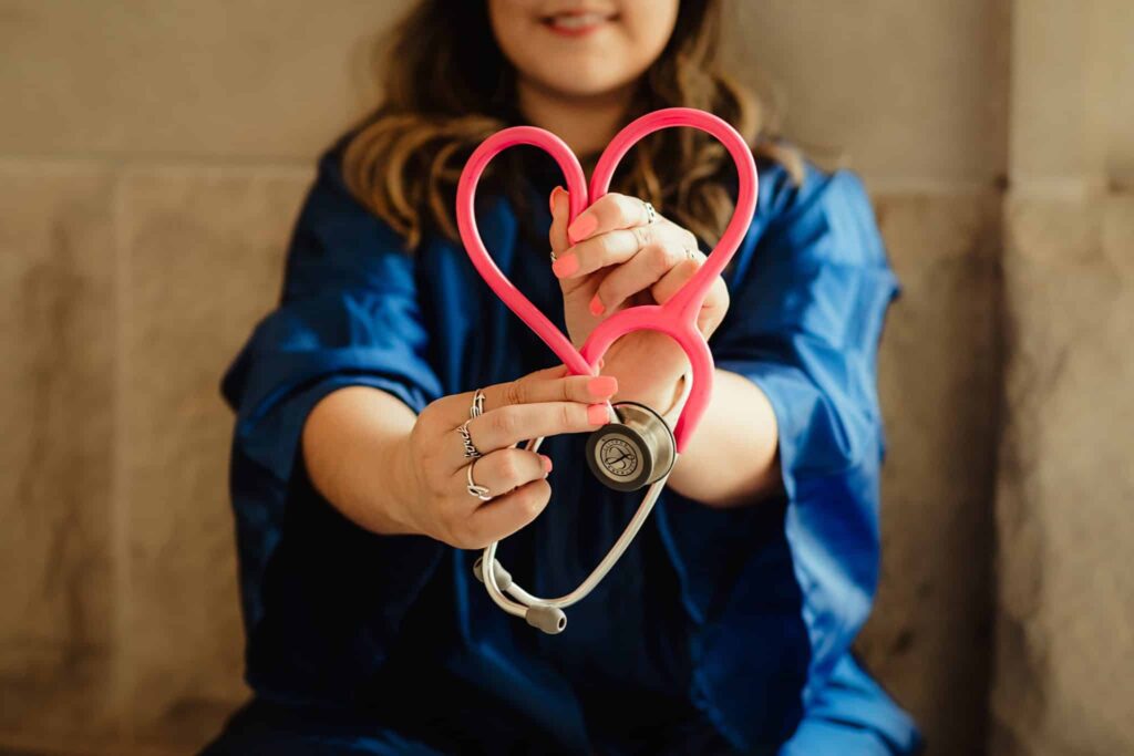A young woman in a graduation robe holding a pink stethoscope in the shape of a heart.