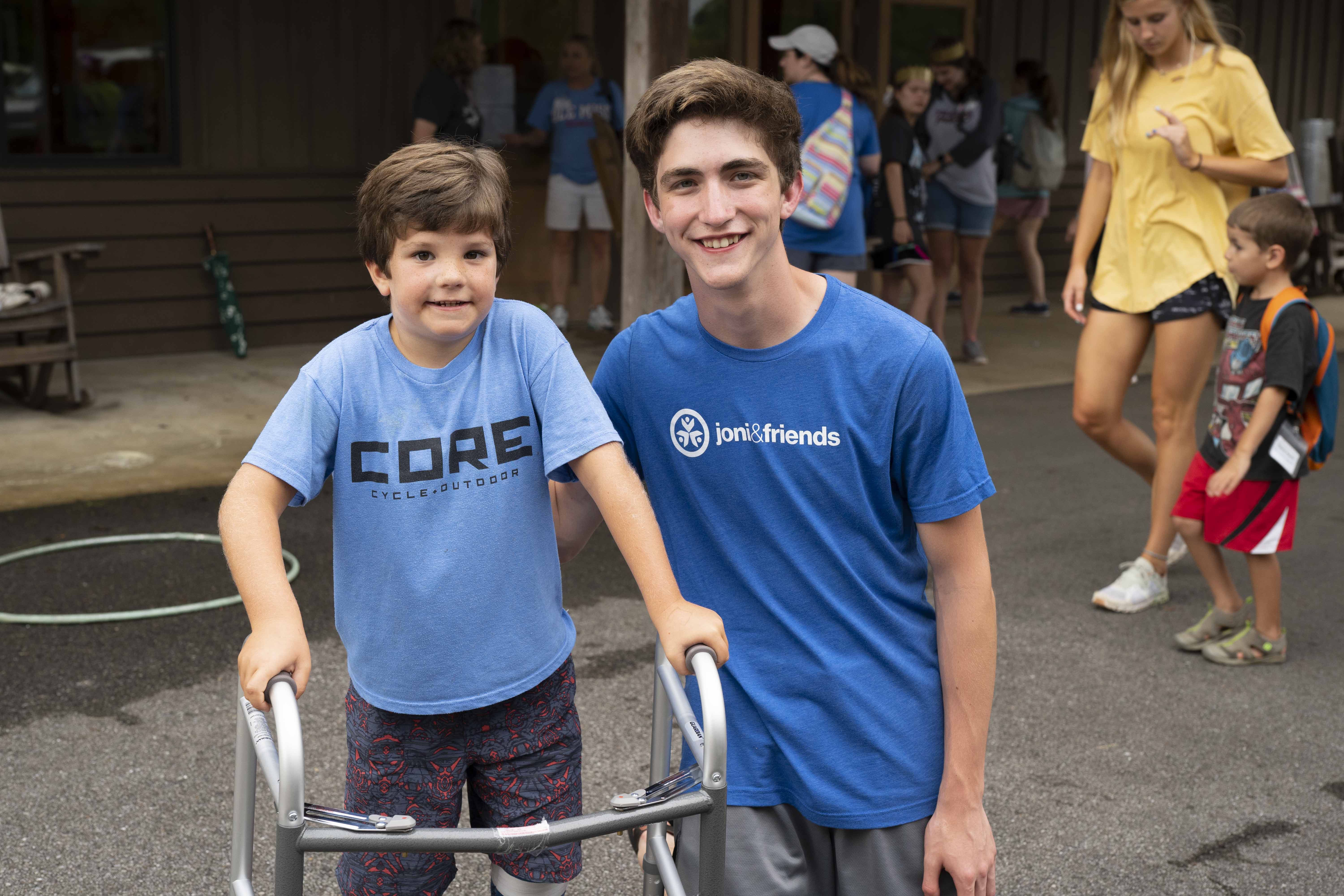 A little boy using a walker with an older boy next to him who's his Family Retreat buddy in a blue Joni and Friends t-shirt. Both smiling at the camera.