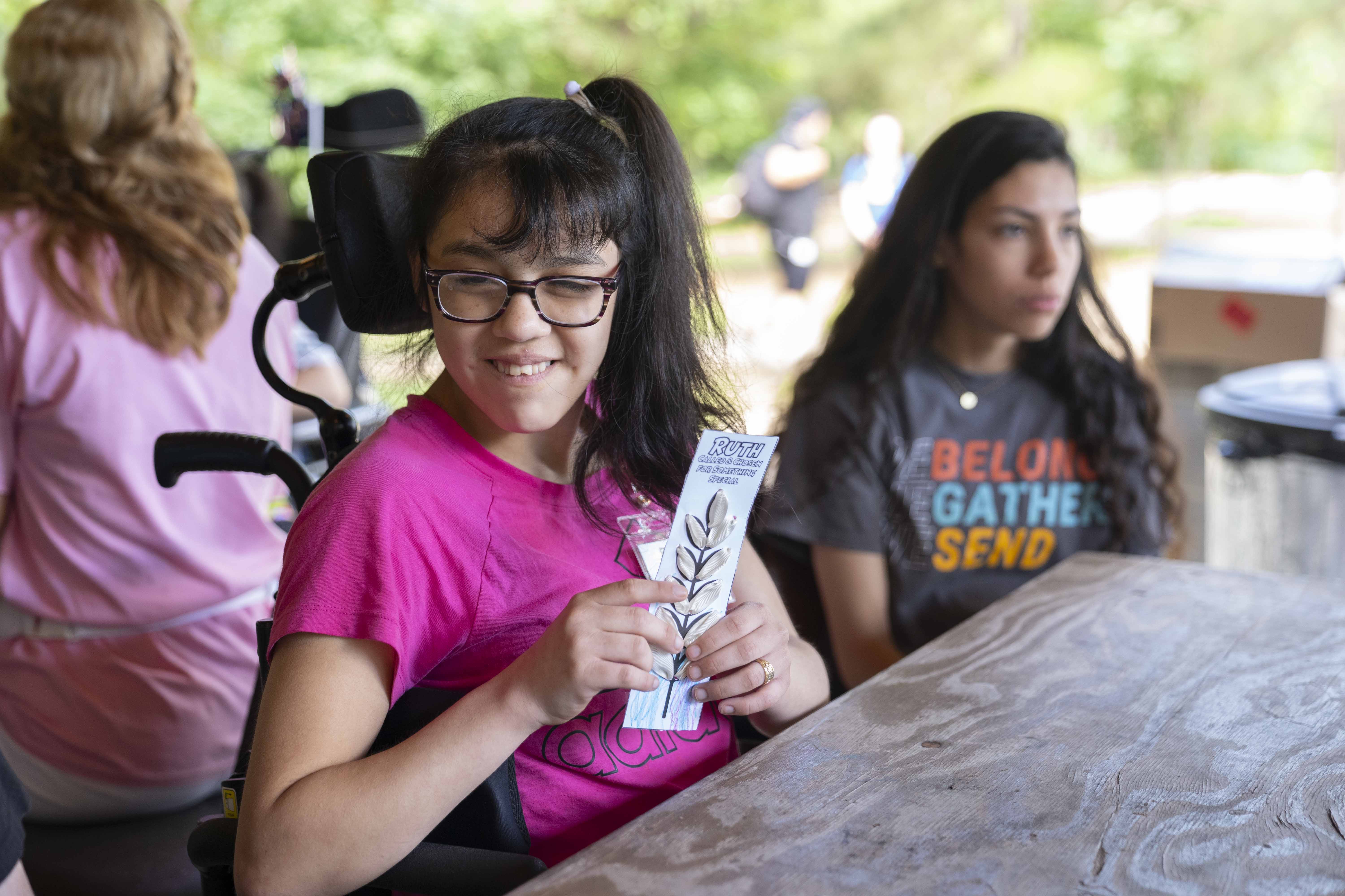 A young girl seated in a wheelchair with a pink shirt on, a side ponytail, and glasses, smiling at the camera.