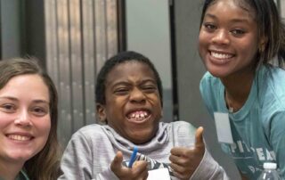 Two volunteers and a young boy are having fun at a respite event. The boy has a huge grin on his face and is giving a double thumbs-up.