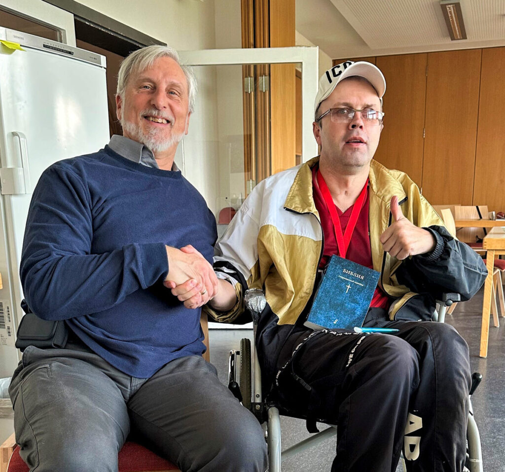 Mark sitting next to a man using a wheelchair with a Bible in his lap in the Ukrainian language. Both smiling at the camera.