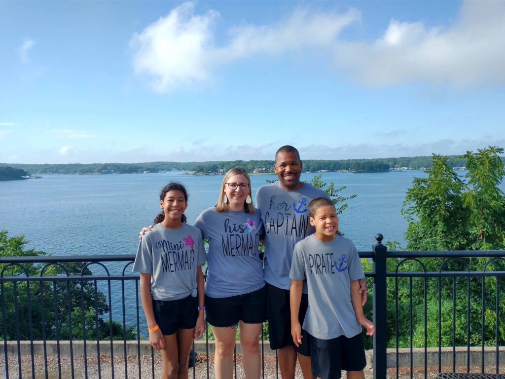 A family photo of Quinn, Mariah, Malakai and Kaiah in front of a lake on a beautiful blue-sky day.