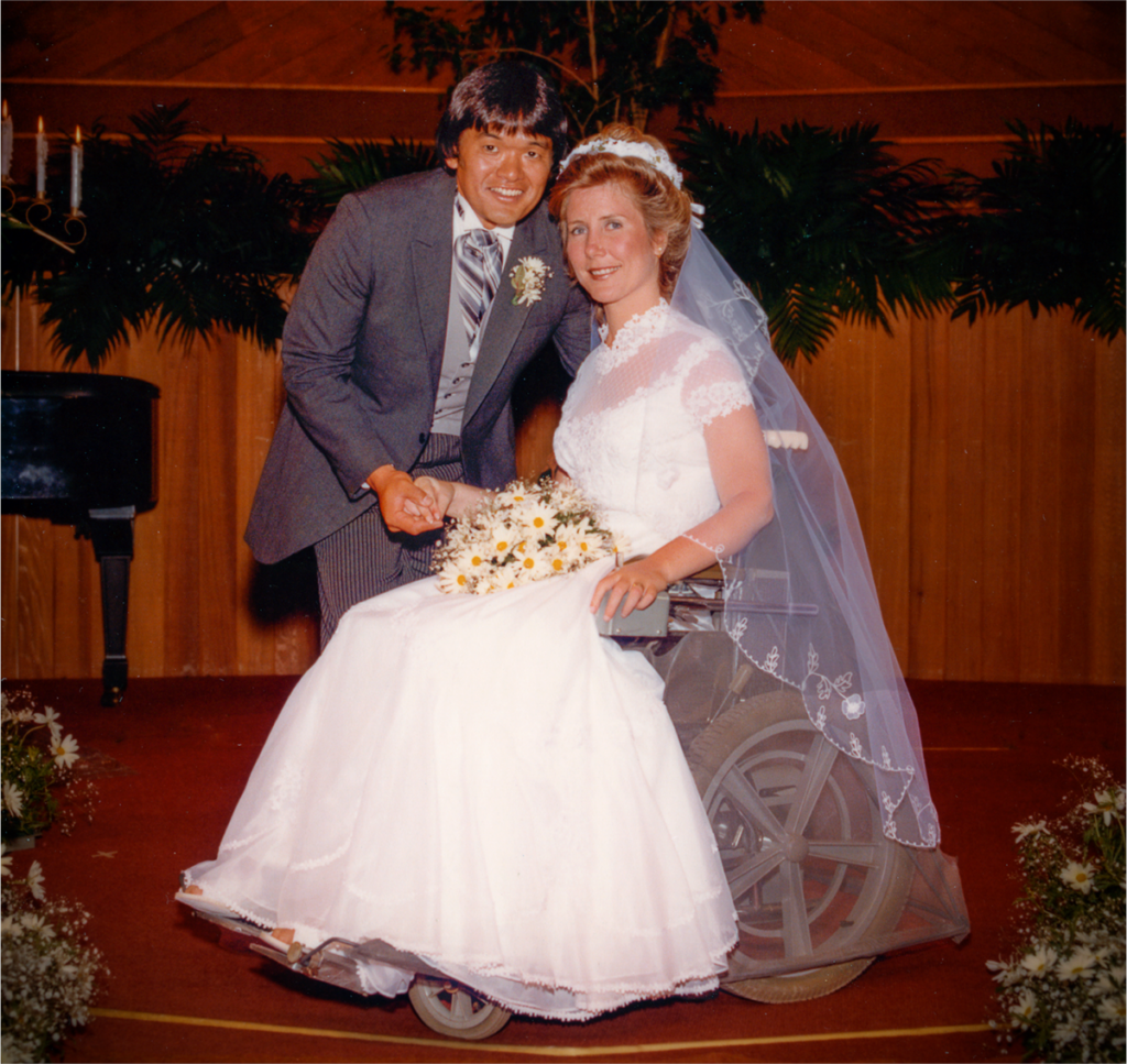 A picture of Joni and Ken on their wedding day at the altar. Joni is seated in her wheelchair with her wedding dress on, Ken leaning over her. Both smiling at the camera.