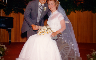 A picture of Joni and Ken on their wedding day at the altar. Joni is seated in her wheelchair with her wedding dress on, Ken leaning over her. Both smiling at the camera.