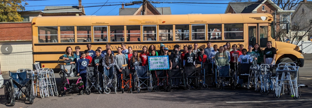 The group of students at Valley Forge, standing in front of a school bus with the wheelchairs they collected, holding a sign that says, "Wheels for the World."