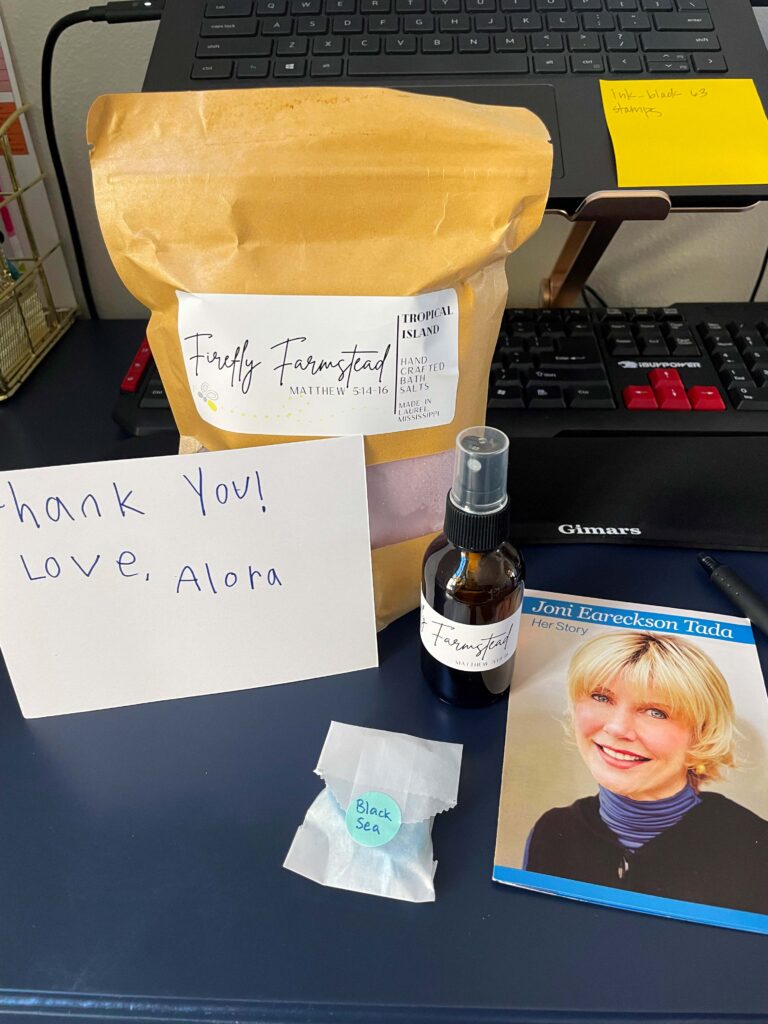 A bag of pink bath salt with a note from Alora, some spray and  black sea soap with a Joni and Friends pamphlet next to it.