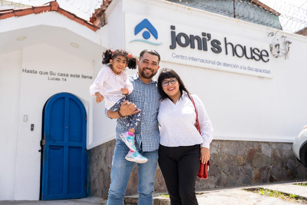 A picture of Sofia being held by her Dad while he is embracing Sofia's mom. The Joni's House El Salvador sign is behind them.
