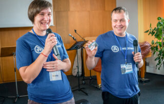 Irena and her husband Oleksiy on stage with their blue Joni and Friends shirts on, smiling as they hold microphones in their hands.