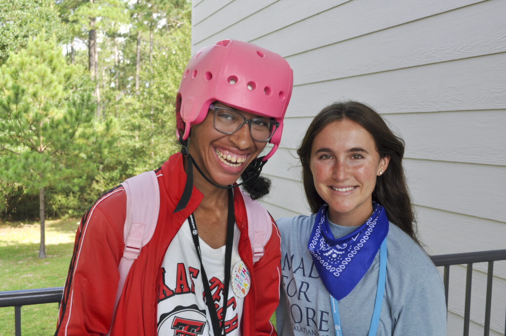 A young woman with a pink protective helmet on who appears to have a disability with her Joni and Friends volunteer next to her with their arm around her. Both smiling at the camera.
