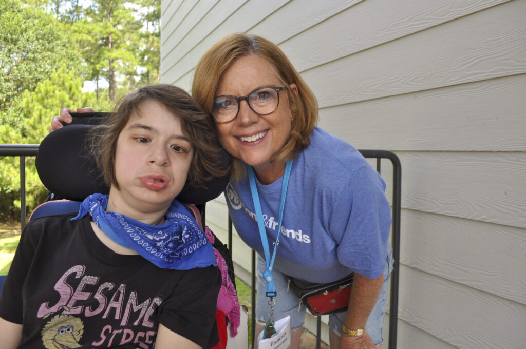 A young woman seated in a wheelchair with her Joni and Friends buddy volunteer standing next to her with their arm wrapped around the back of her chair. Both smiling at the camera.
