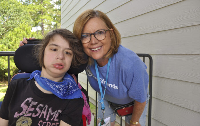 A young woman seated in a wheelchair with her Joni and Friends buddy volunteer standing next to her with her arm wrapped around the back of her chair. Both smiling at the camera.