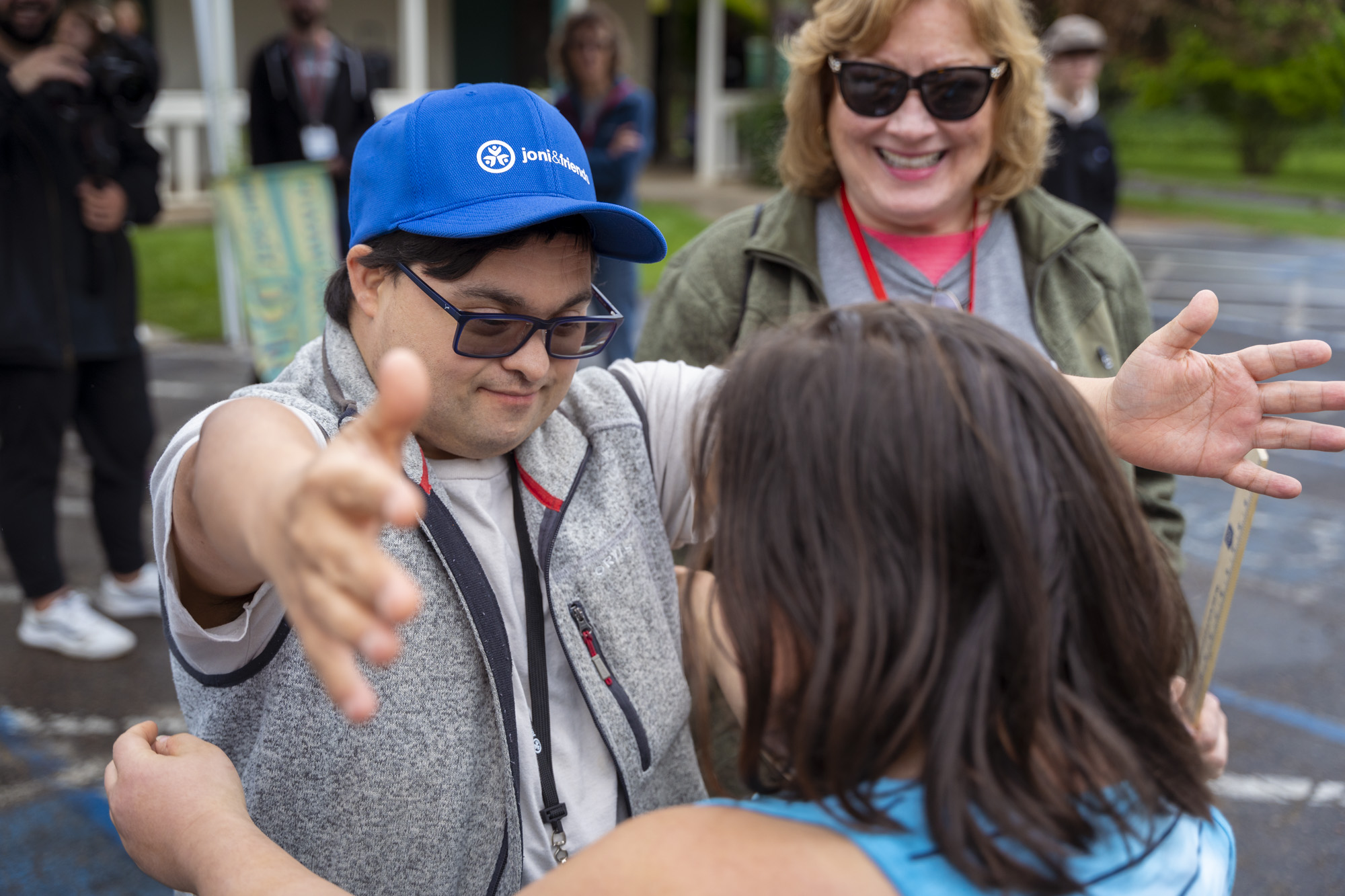 A picture of a young man with Down Syndrome wearing a Joni and Friends hat as he goes in to hug a friend. A woman is standing behind him and smiling.