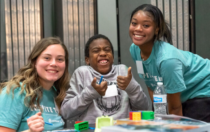 A picture of John at half-time respite with his two Joni and Friends buddy volunteers on either side of him. He is smiling wide at the camera with his two thumbs up.