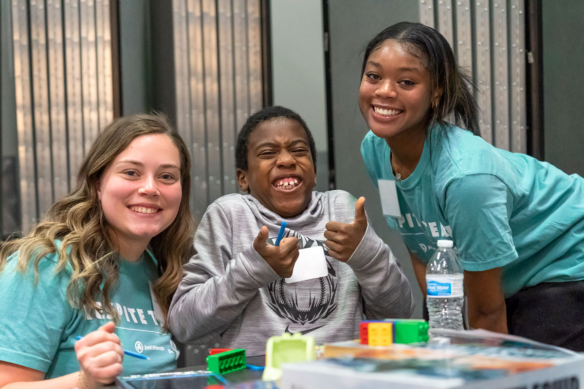 A picture of John at half-time respite with his two Joni and Friends buddy volunteers on either side of him. He is smiling wide at the camera with his two thumbs up.