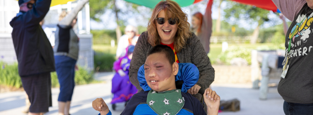 A young boy in a wheelchair is being pushed under a billowing parachute. The boy is smiling and his hands are raised. He is experiencing joy and love at Family Retreat.